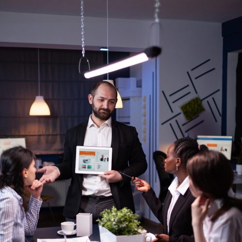 Business entrepreneur man presenting company statistics using tablet for financial presentation. Diverse multi-ethnic businesspeople brainstorming strategy working in office meeting room late at night